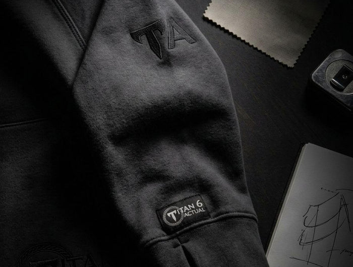 A dark charcoal gray hoodie laid flat on a dark surface, featuring tonal black embroidered logos on the sleeve and a visible 'Titan 6' brand patch near the cuff. Styled flat-lay props include fabric swatches and a sketchbook in the background.