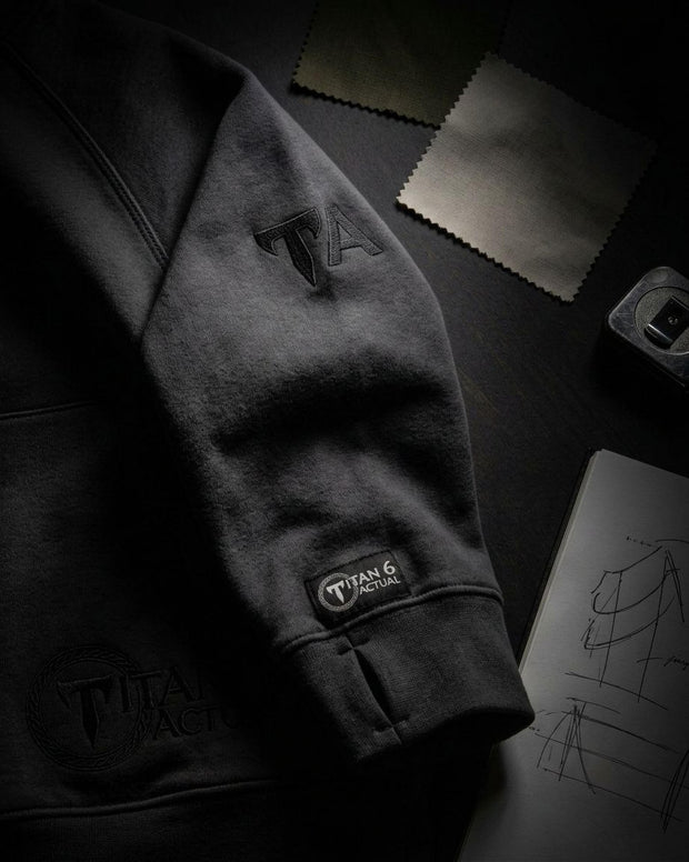 A dark charcoal gray hoodie laid flat on a dark surface, featuring tonal black embroidered logos on the sleeve and a visible 'Titan 6' brand patch near the cuff. Styled flat-lay props include fabric swatches and a sketchbook in the background.