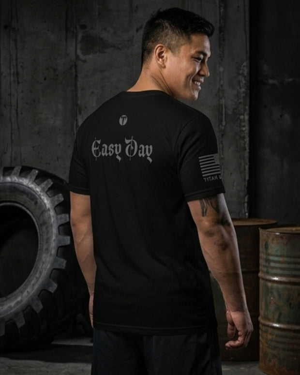A man wearing a black t-Titan 6 Endure T-shirt in with 'Easy Day' printed in white Gothic font on the back, standing in a dark industrial setting with a tire and metal barrels.