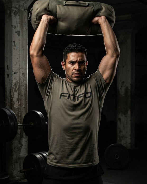 A man in a gym wearing a fitted army green Titan 6 Endure T-shirt with 'FITFO' printed on the chest, lifting a sandbag overhead.