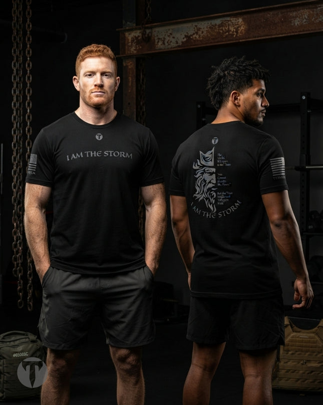 Two men modeling black Titan 6 Endure T-shirt with the Storm design in a dark gym setting. The front view shows a man with red hair wearing a black t-shirt with a white 'I AM THE STORM' graphic and a small Icon logo.