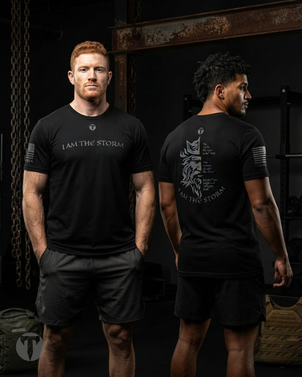 Two men modeling black Titan 6 Endure T-shirt with the Storm design in a dark gym setting. The front view shows a man with red hair wearing a black t-shirt with a white 'I AM THE STORM' graphic and a small Icon logo.