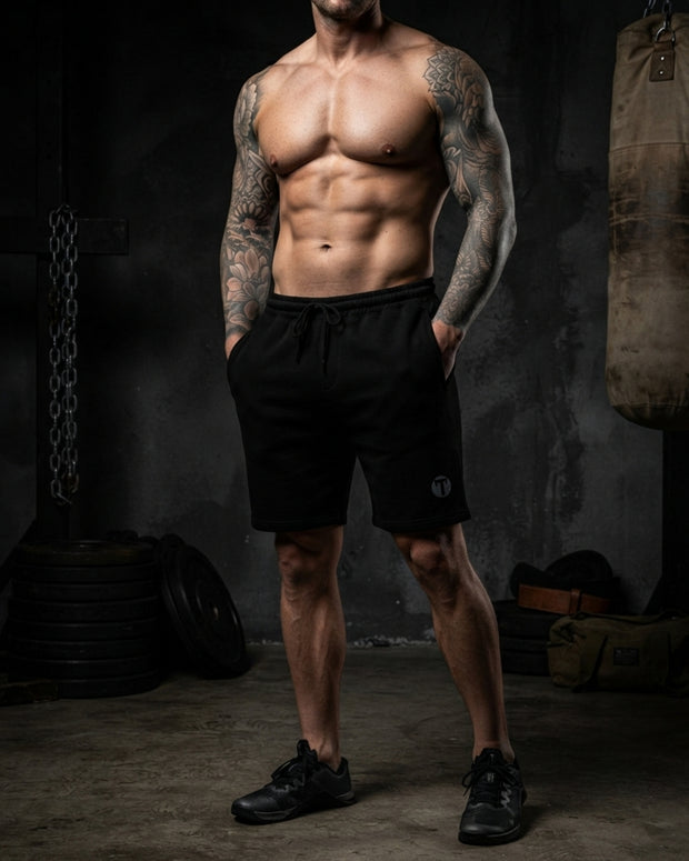 A muscular man with tattoos stands in a dimly lit gym wearing black Titan 6 Outpost shorts with the light Icon Logo and black athletic shoes. Barring, weights, and a punching bag are visible in the background.