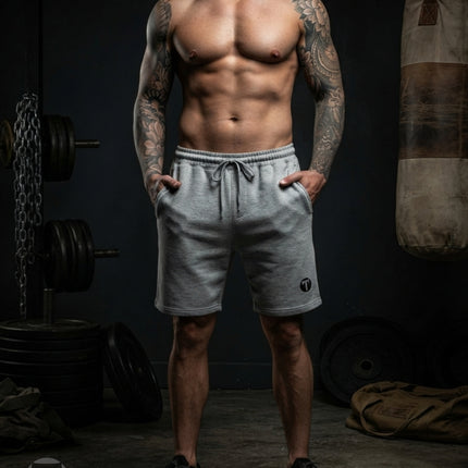A muscular man with tattoos stands in a dimly lit gym wearing light grey Titan 6 Outpost shorts with the dark Icon Logo and black athletic shoes. Barring, weights, and a punching bag are visible in the background.