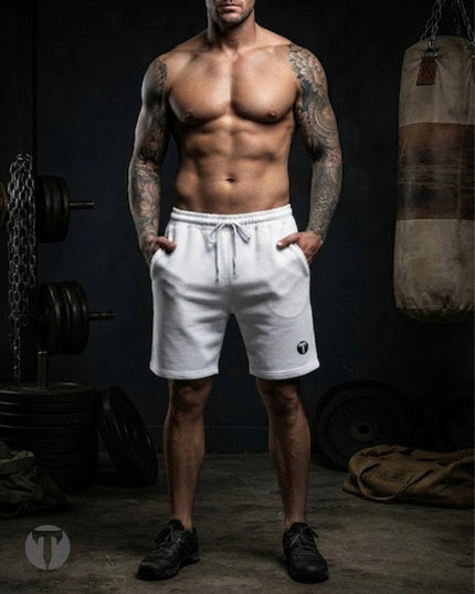 A muscular man with tattoos stands in a dimly lit gym wearing white Titan 6 Outpost shorts with the dark Icon Logo and black athletic shoes. Barring, weights, and a punching bag are visible in the background.