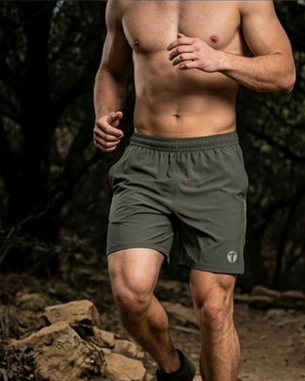 A shirtless male runner wearing army green Titan 6 Stamina Drawstring 5 Inch Linerless shorts with a small light Icon logo on the lower left leg, captured mid-stride in a dark forest setting.