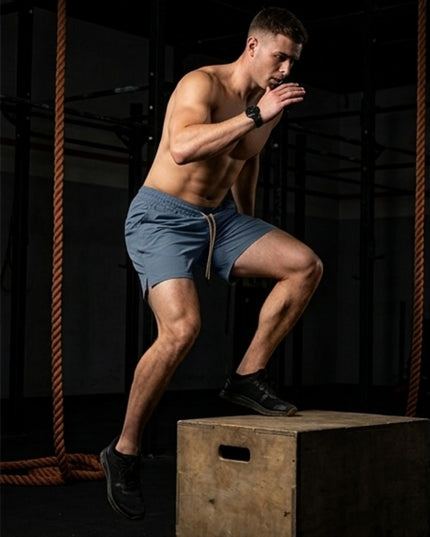 A shirtless man in grey blue Titan 6 Stamina Drawstring 5 Inch Linerless shorts and black sneakers performs a box jump on a wooden plyo box in a dark gym setting with ropes.