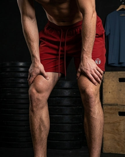 A man in a gym wearing red Titan 6 Stamina Drawstring 5 Inch Linerless shorts, leaning forward with hands on his knees. The background features stacked black weight plates and wooden boxes.