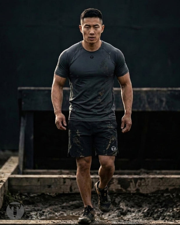 A muscular man wearing a dark gray Titan 6 Strike Raglan T-shirt with the dark Icon Logo and matching shorts walks through muddy terrain in an outdoor setting.