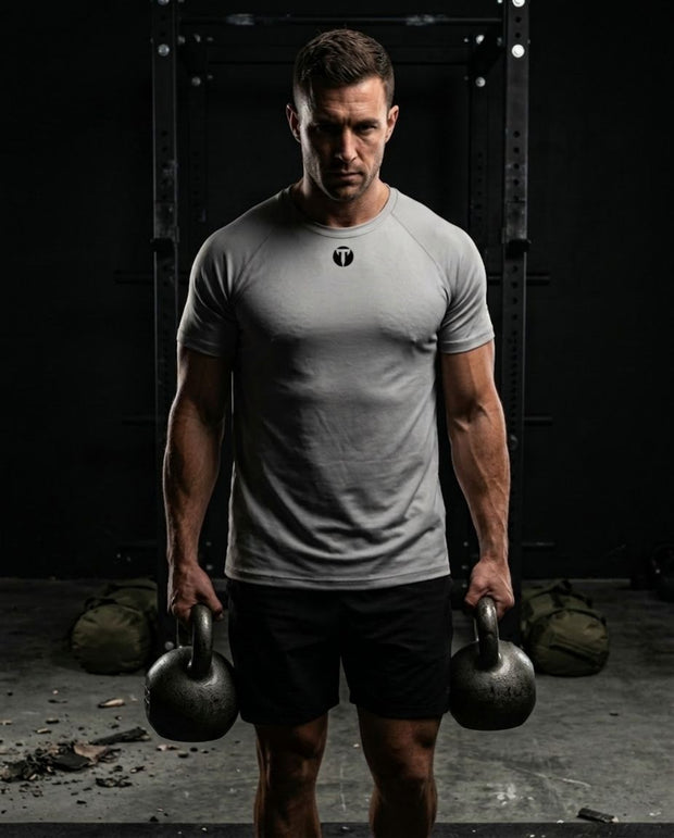 A muscular man in a gym setting wearing a Titan 6 Strike Raglan T-shirt in light gray with the dark Icon logo on the chest, paired with black shorts, holding two kettlebells.