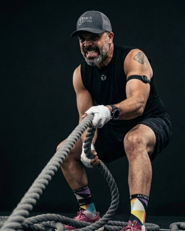 Titan 6 Athlete, Jon Ross-Wiley, in athletic wear, including a black Titan 6 Vanguard Muscle Shirt with the light Icon logo, black shorts, and a dark gray Titan 6 cap, is shown from the waist up while pulling a thick battle rope against a dark studio background. He is wearing white gloves, a black armband monitor, and colorful athletic socks.