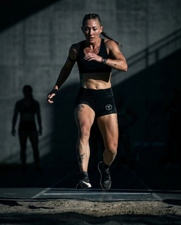 Titan 6 Athlete Kelly Sullivan in a black Glide sports bra and black Glide shorts with the Titan's Shield Elite logo on the left leg, running during a CrossFit workout in a dark concrete gym environment.