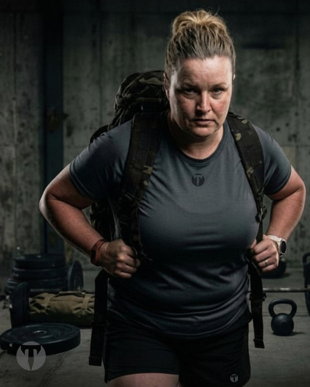 Titan 6 Athlete Nicole Kelly in a gym setting wearing a dark gray Strike Raglan t-shirt, black Stamina Drawstring shorts, and a large black Frontline Athletic Ruck, with a weight plate and kettle bells visible in the background.