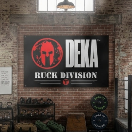 DEKA Ruck Division Gym Flag featuring a red Spartan helmet logo, the Titan 6 Actual Icon Logo, and bold text, displayed on a brick wall inside a training facility with bumper plates, kettlebells, and gear.