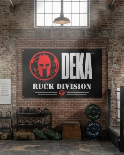 DEKA Ruck Division Gym Flag featuring a red Spartan helmet logo, the Titan 6 Actual Icon Logo, and bold text, displayed on a brick wall inside a training facility with bumper plates, kettlebells, and gear.
