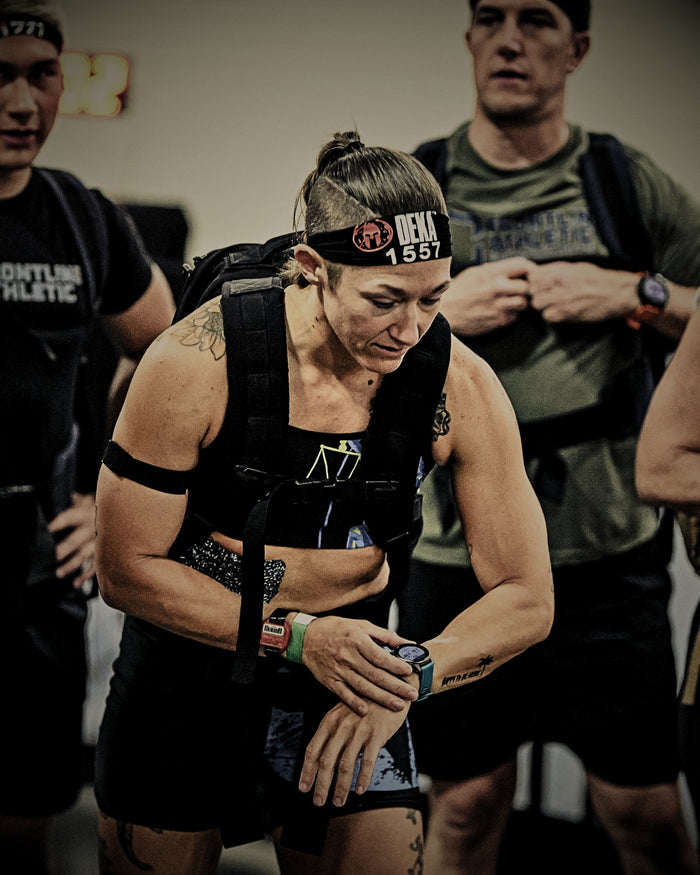 A female Titan 6 Athlete checking her watch at the Spartan DEKA FIT Ruck start line, wearing athletic apparel and a weighted rucksack.