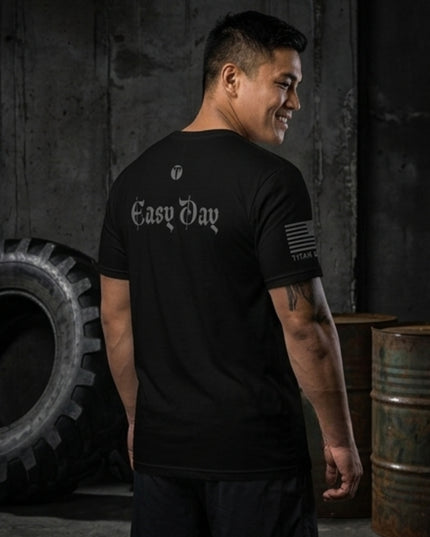 A man wearing a black t-shirt with 'Easy Day' printed in white Gothic font on the back, standing in a dark industrial setting with a tire and metal barrels.