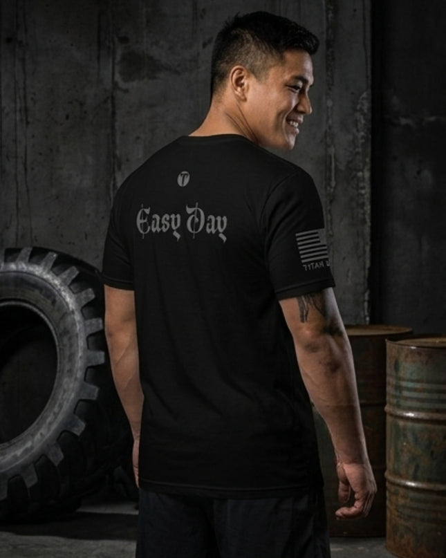 A man wearing a black t-shirt with 'Easy Day' printed in white Gothic font on the back, standing in a dark industrial setting with a tire and metal barrels.