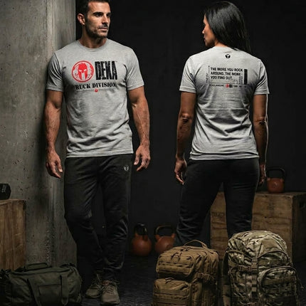 Male and female athletes wearing gray Spartan DEKA Ruck Division graphic t-shirts alongside tactical rucksacks and kettlebells in a training facility.
