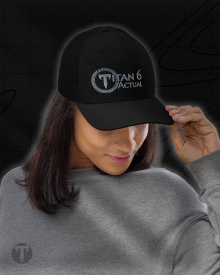 A woman wearing a black baseball cap with a white 'TITAN 6 ACTUAL' logo on the front, paired with a gray sweatshirt against a dark graphic background.