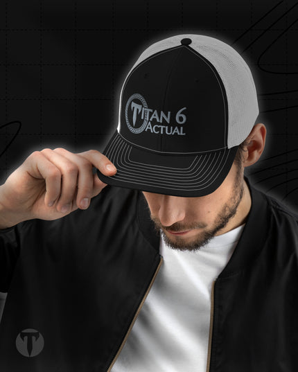 A man wearing a black and white trucker hat with 'TITAN 6 ACTUAL' printed on the front, holding the brim down over his eyes. The background is a dark, textured studio setting.