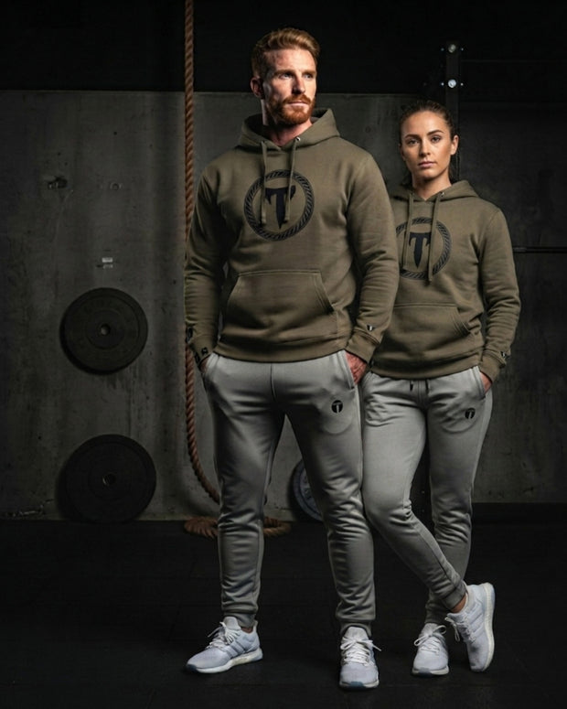 A athletic man and woman standing together in a gym setting wearing matching olive green Titan 6 hoodies with the dark Elite logo on the chest and light grey joggers featuring the dark Icon logo on the thigh.