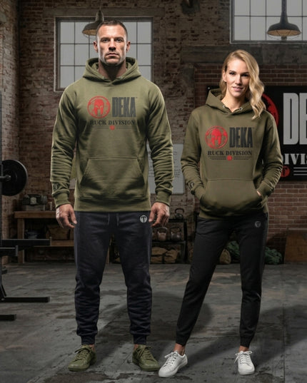 A man and a woman modeling matching olive green Titan 6 Citadel  hoodies with a red and black Spartan 'DEKA RUCK DIVISION' and red Titan 6 Icon logo on the chest, set in a rustic industrial loft with brick walls and gym equipment.