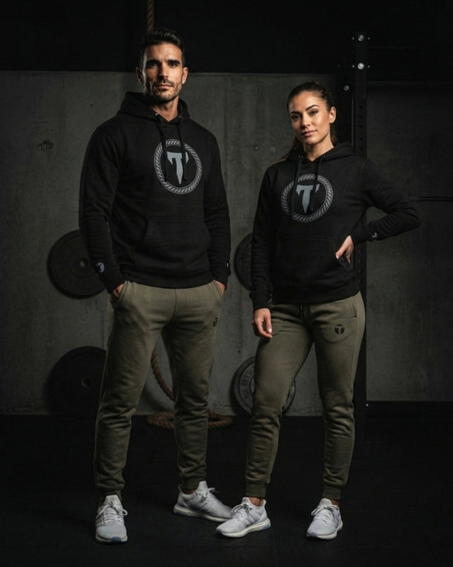 A male and female athlete posing in a gym setting wearing matching black Titan 6 Citadel Pullover Hoodies with the light Elite logo and olive green joggers featuring the dark Icon logo on the thigh.