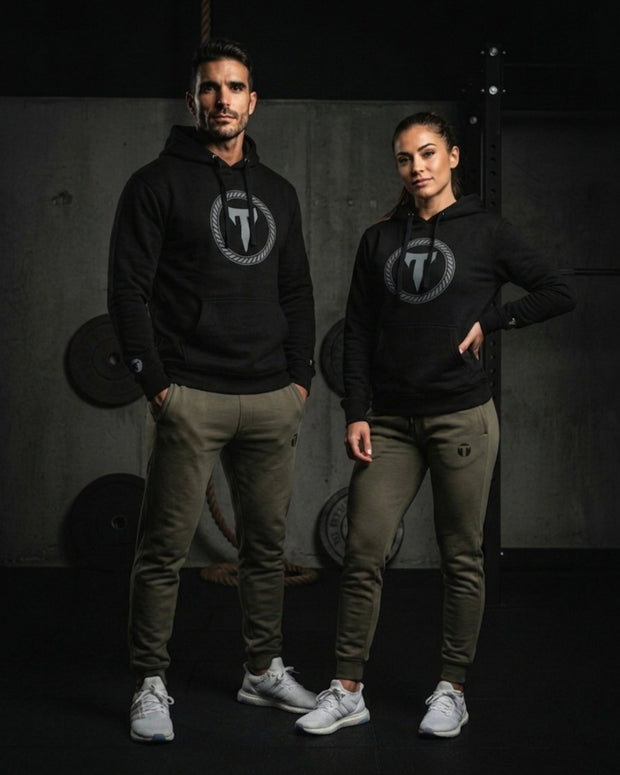 A male and female athlete posing in a gym setting wearing matching black Titan 6 Citadel Pullover Hoodies with the light Elite logo and olive green joggers featuring the dark Icon logo on the thigh.