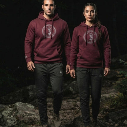 Man and woman wearing a maroon Titan 6 Citadel Pullover Hoodie featuring the circular Zeus emblem, paired with dark grey joggers while walking on a rocky outdoor trail.