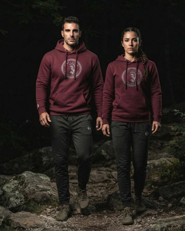 Man and woman wearing a maroon Titan 6 Citadel Pullover Hoodie featuring the circular Zeus emblem, paired with dark grey joggers while walking on a rocky outdoor trail.