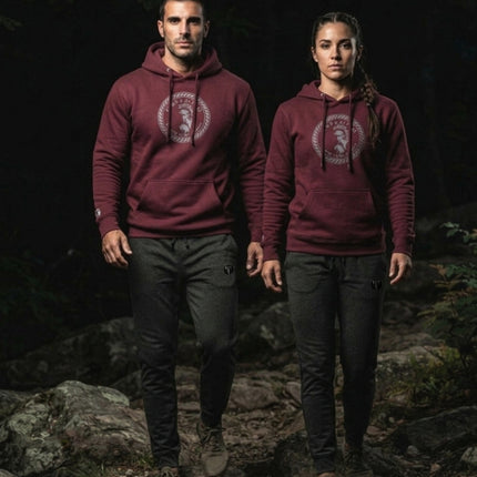A man and a woman standing on a rocky forest path, both wearing matching maroon Titan 6 Citadel hoodies with the Zeus design on the chest and dark Charcoal Titan 6 Garrison Joggers featuring the Icon logo.