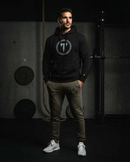 Athletic man wearing dark green Titan 6 Garrison Joggers with a subtle "T" logo on the left thigh, paired with a black Titan 6 pullover hoodie and white sneakers in a gym setting.