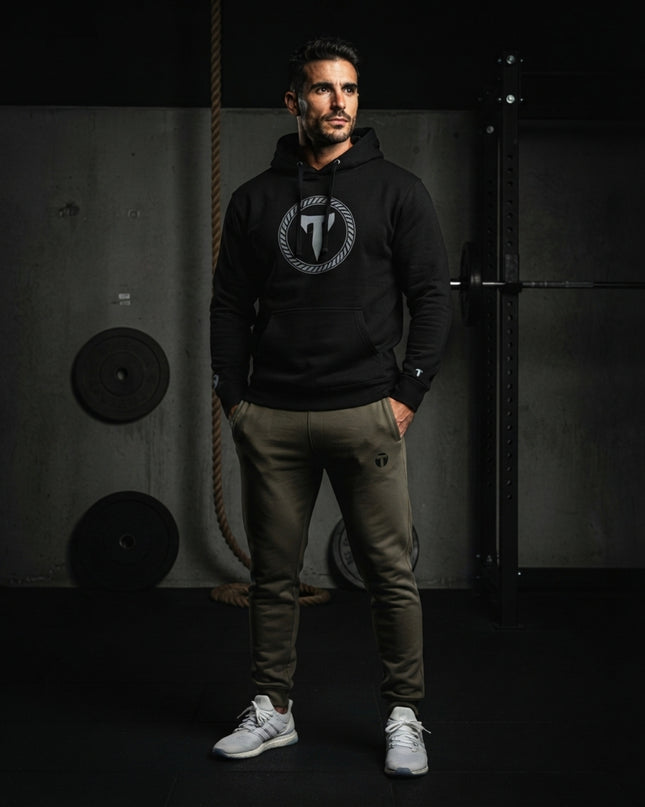 Athletic man wearing dark green Titan 6 Garrison Joggers with a subtle "T" logo on the left thigh, paired with a black Titan 6 pullover hoodie and white sneakers in a gym setting.