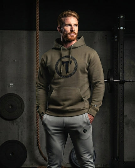 Athletic man wearing Athletic Grey Titan 6 Garrison Joggers with a subtle "T" logo on the left thigh, paired with a olive green Titan 6 pullover hoodie and white sneakers in a gym setting.