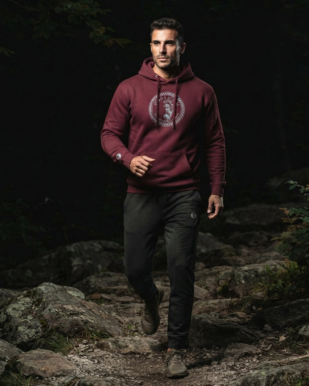 Athletic man wearing dark Charcoal Heather Titan 6 Garrison Joggers with a subtle "T" logo on the left thigh, paired with a maroon Titan 6 pullover Zeus hoodie and white sneakers in a gym setting.