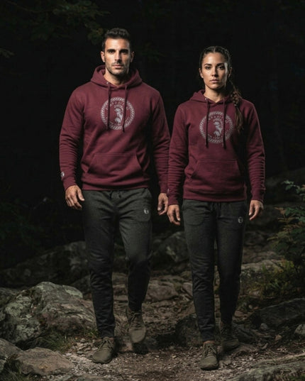 Athletic man and woman wearing dark Charcoal Heather Titan 6 Garrison Joggers featuring the light Titan 6 Icon logo on their left thigh, paired with a maroon Titan 6 pullover Zeus hoodie and white sneakers in a gym setting.