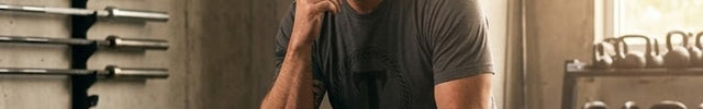 Titan 6 Actual owner and founder, Lou Greco, wearing a grey Titan 6 graphic t-shirt sitting thoughtfully on a wooden plyo box in a gym, with hand under chin surrounded by barbells and kettlebells.