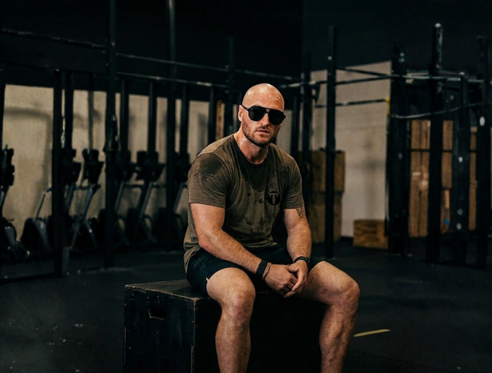 Titan 6 Athlete, Zack Wisnowski, wearing sunglasses and a sweat-stained olive green Titan 6 Nomad tee sitting on a wooden plyo box in a gym.