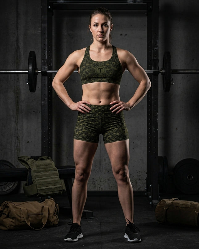 Athletic woman wearing the Titan 6 Glide Sports Bra and Glide Shorts set in olive drab Golgotha skull print, standing in a gym in front of a squat rack with workout gear nearby.