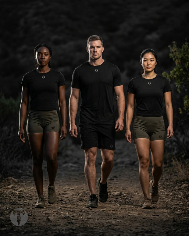 Three fitness models walking side-by-side on a dark dirt trail, wearing matching black Titan 6 Breeze t-shirts.  The women are wearing olive green Titan 6 Agility shorts and the man is wearing black Titan 6 Stamina Drawstring 5 Inch linerless shorts.