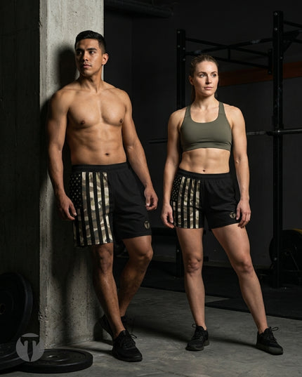 A shirtless man and a woman standing in a gym, both wearing matching Titan 6 Endure 6.5 inch linerless shorts in the Shadow Patriot pattern. The man wears black athletic shoes, and the woman wears black athletic shoes with a green sports bra.