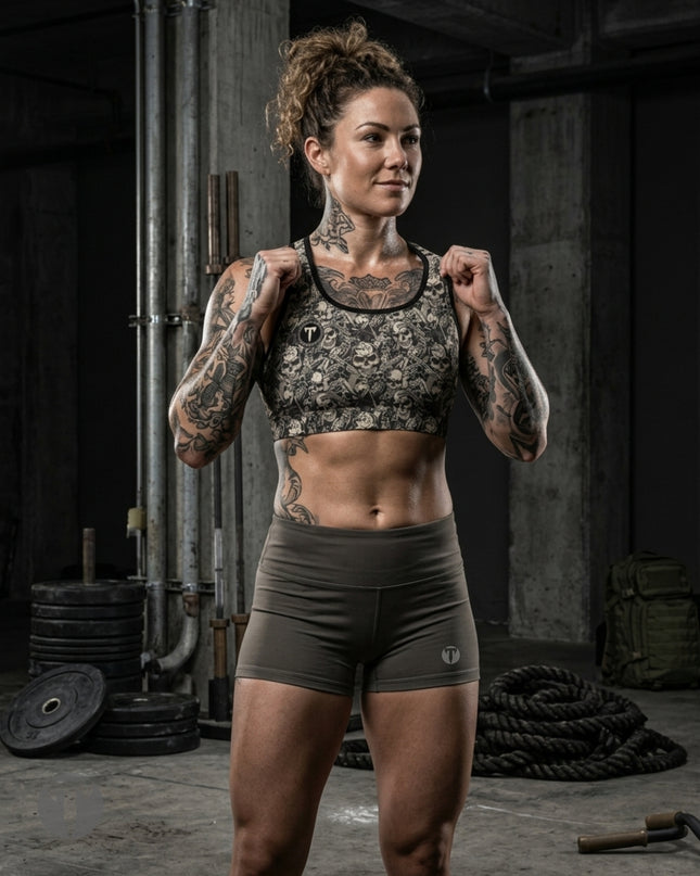 A tattooed woman stands in a gritty gym setting wearing a Titan 6 Glide Sports Bra in Outlaw pattern and matching dark olive green athletic shorts.