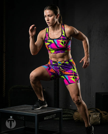 A woman in a vibrant, multicolor geometric print Titan 6 Glide Sports Bra and matching Glide shorts Zack Wiz set performs a step up on a black plyo box in a dark gym setting.