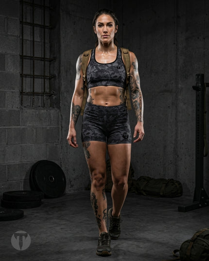 A woman with tattoos walks forward in a dark gym setting wearing a Titan 6 Glide Sports Bra in Twilight Camo and matching Glide shorts, with a tan backpack on her shoulders.