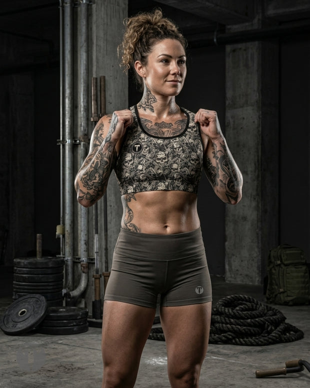A tattooed woman stands in a gritty gym setting wearing a Women's Titan 6 Glide Sports Bra in an Outlaw patterned sports bra and matching dark olive green Women's Titan 6 Glide Shorts.