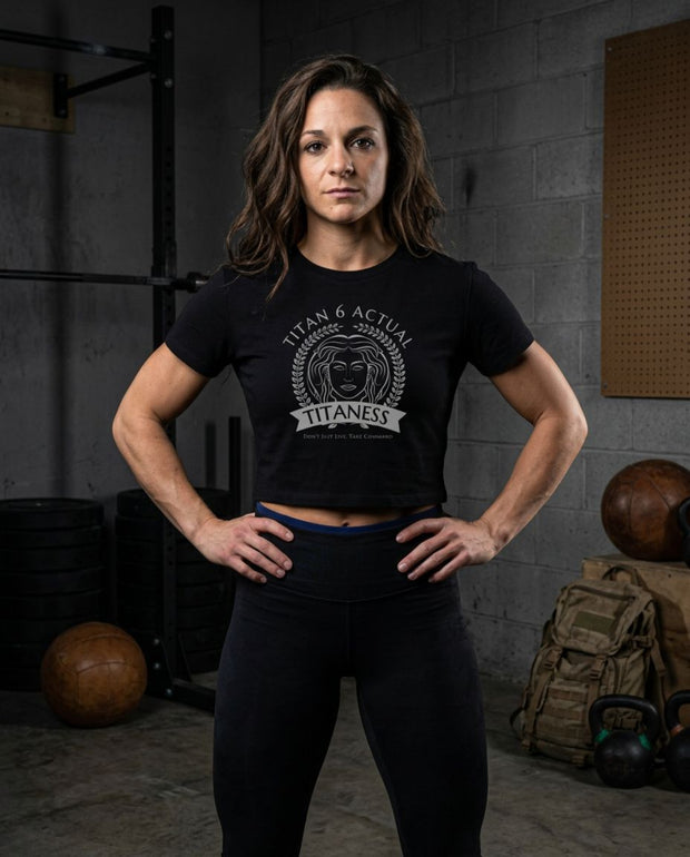 Titan 6 Athlete, Silvana Fazzalari, stands in a gym wearing a black Titan 6 Valor Crop T-shirt with a white circular graphic that reads 'TITAN 6 ACTUAL TITANESS' and black leggings.